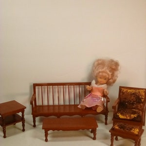 May include: A dollhouse furniture set with a brown couch, two end tables, a coffee table, and a chair with a footstool. The furniture is made of wood and has a rustic look. A doll is sitting on the couch.
