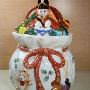May include: White ceramic Christmas cookie jar shaped like a Santa sack with a toy soldier, teddy bear, and train on the front. The lid features a toy soldier holding a drum.