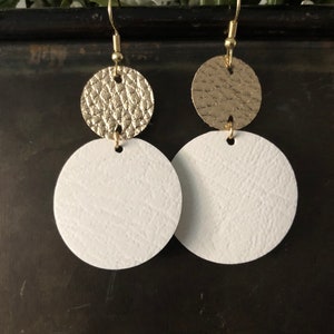 Puede incluir: Un par de pendientes de cuero dorado y blanco. Los pendientes están hechos de dos círculos, un círculo dorado más pequeño en la parte superior de un círculo blanco más grande.