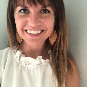 Op de afbeelding: Een vrouw met lang bruin haar die een wit geplooid topje en kurk oorbellen in de vorm van bladeren draagt.
