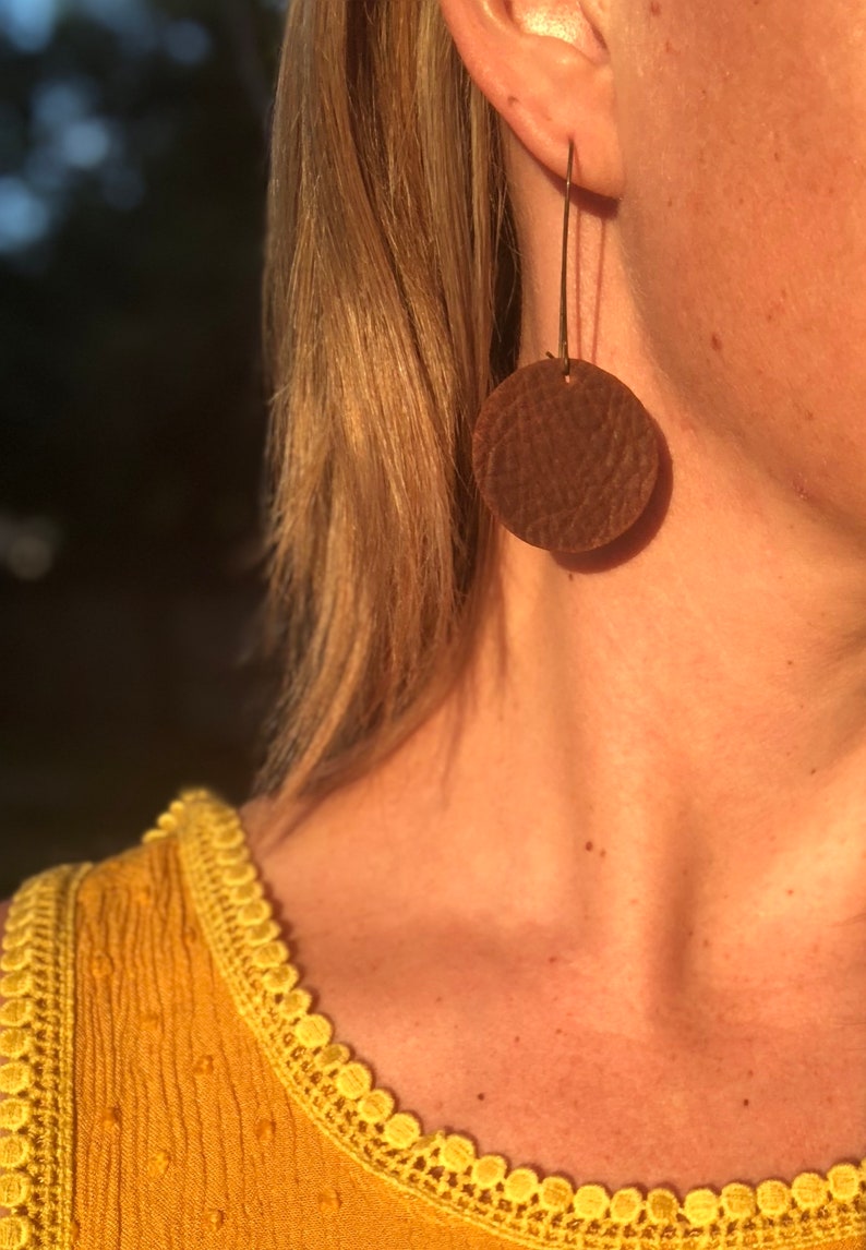 May include: Brown leather circle earrings with a textured surface hanging from gold-toned hooks.