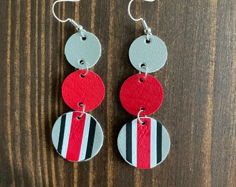 May include: A pair of dangle earrings with a silver hook and three stacked, circular elements. The top and middle circles are solid silver and red, respectively. The bottom circle is silver with red and black stripes.