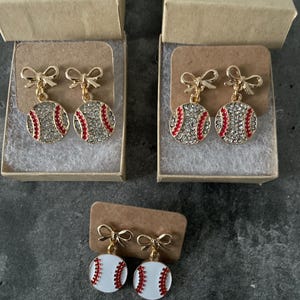 Könnte beinhalten: Ohrringe zum Thema Baseball. Zwei Paar Ohrringe zeigen Basebälle, die mit klaren Strasssteinen und roten Nähten besetzt sind und an goldfarbenen Schleifen hängen. Ein drittes Paar hat weiße Basebälle mit roten Nähten. Alle werden in kleinen, offenen Pappschachteln präsentiert.