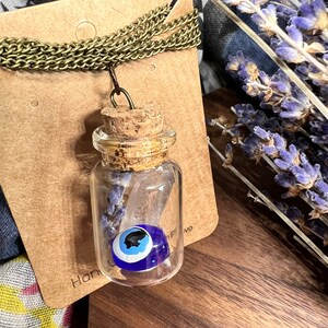 May include: A glass bottle necklace with a cork stopper and a blue evil eye charm inside. The bottle is filled with dried lavender. The necklace is on a brown card with the word "Hand" visible.