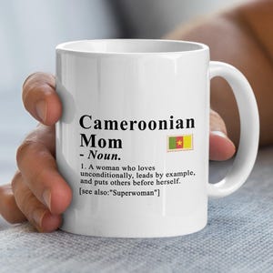 May include: A white ceramic mug featuring black printed text that reads: Cameroonian Mom - Noun. 1. A woman who loves unconditionally, leads by example, and puts others before herself. [see also: "Superwoman"]. A small rectangular Cameroonian flag is included.