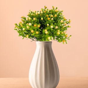 Może przedstawiać: Biały, żebrowany ceramiczny wazon z małym układem zielonych i żółtych sztucznych kwiatów. Wazon stoi na drewnianej powierzchni na tle brzoskwiniowego tła, tworząc prostą, elegancką ekspozycję.