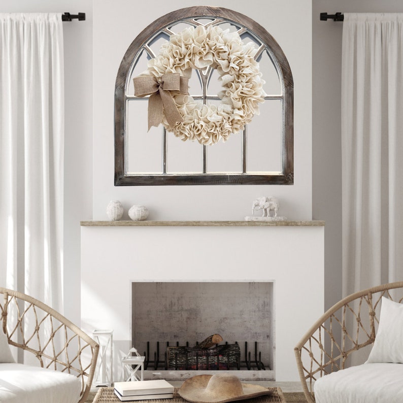 May include: A white and brown farmhouse style wreath with a burlap bow hanging on a wooden arched window frame above a white fireplace.