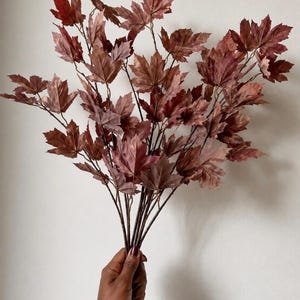 May include: A bundle of artificial maple branches featuring deep burgundy and dark brown leaves. The foliage is arranged on thin, dark brown stems that branch out to display numerous serrated leaves with visible veins, mimicking the appearance of autumn.