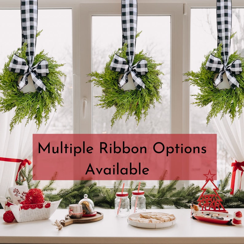 Op de afbeelding: Drie zwart-wit geblokte lintkransen hangen in een raam met de tekst "Multiple Ribbon Options Available". De kransen zijn gemaakt van groen blad en hangen aan de bovenkant van het raam. De tekst staat in een rode doos.