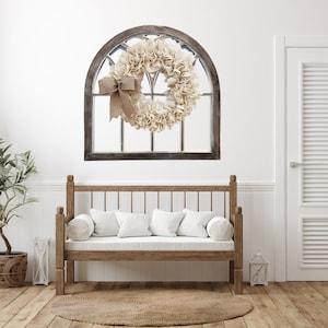 May include: A rustic wooden arched window frame with a burlap and cream fabric wreath hanging on a white wall. A wooden bench with white cushions sits in front of the window.