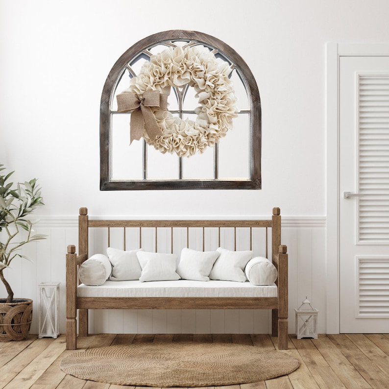 May include: A rustic wooden arched window frame with a burlap and cream fabric wreath hanging on a white wall. A wooden bench with white cushions sits in front of the window.
