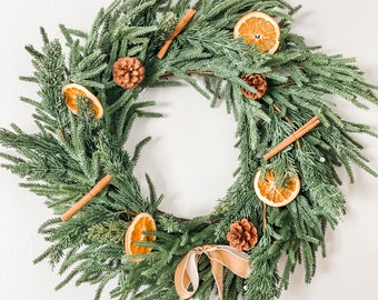 May include: A green evergreen wreath decorated with dried orange slices, cinnamon sticks, and pine cones. A brown ribbon hangs from the bottom of the wreath.