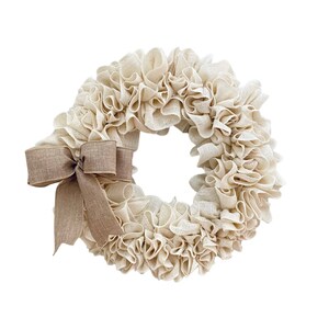 May include: A cream-colored burlap wreath with a brown burlap bow. The wreath is made of fabric and has a ruffled texture.