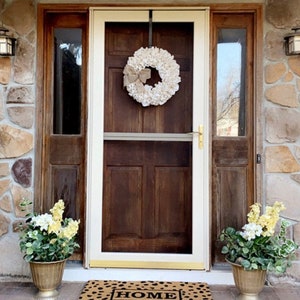 May include: A white burlap wreath with a brown bow hangs on a brown wooden door with a white frame. The door has a screen door and a glass panel on either side. There are two potted plants with yellow flowers on either side of the door. The doormat says "HOME".