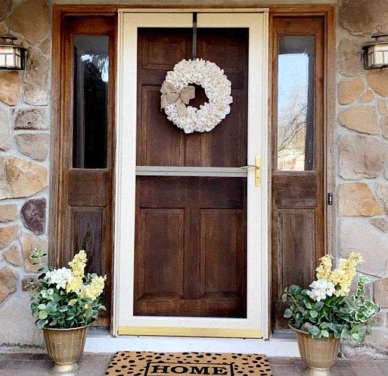 May include: A white burlap wreath with a brown bow hangs on a brown wooden door with a white frame. The door has a screen door and a glass panel on either side. There are two potted plants with yellow flowers on either side of the door. The doormat says "HOME".