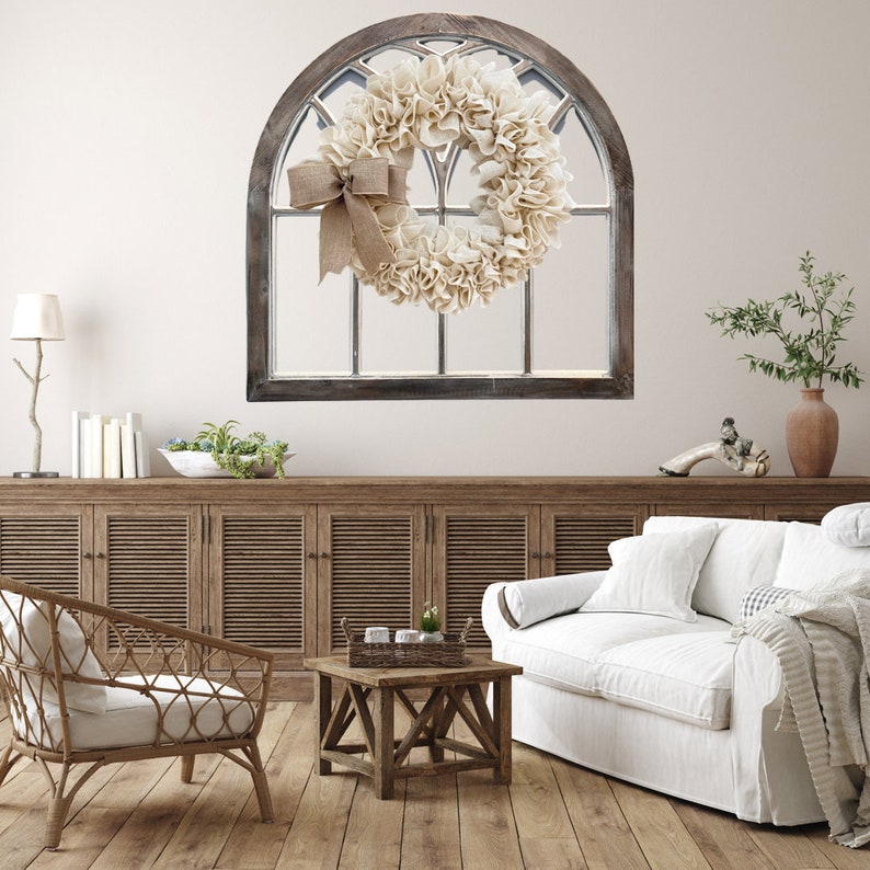 May include: A white farmhouse style wreath with a burlap bow hangs on a distressed wood arched window frame. The wreath is made of fabric and has a rustic look. The window frame is mounted on a white wall.