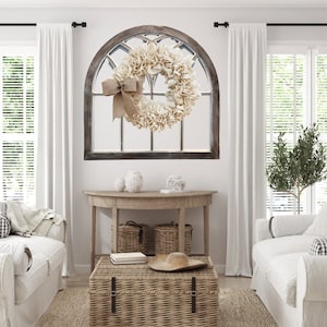 May include: A white farmhouse style wreath with a burlap bow hanging on a wooden arched window frame. The wreath is made of fabric and has a rustic look. The window frame is painted a dark brown color and has a distressed finish.