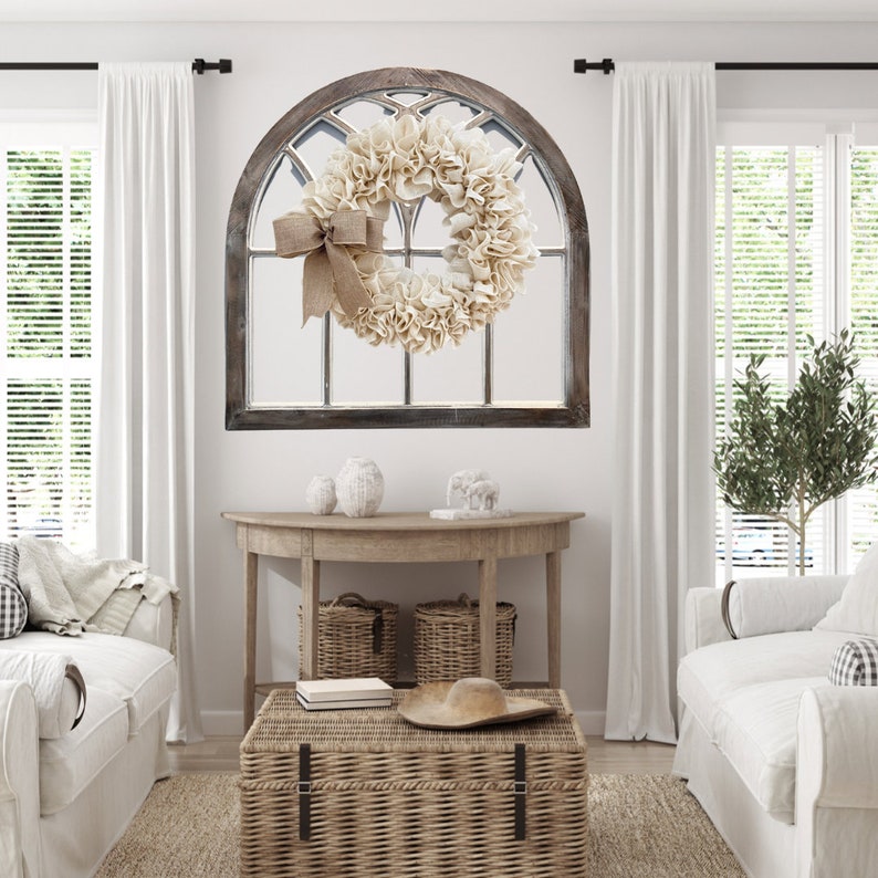 May include: A white farmhouse style wreath with a burlap bow hanging on a wooden arched window frame. The wreath is made of fabric and has a rustic look. The window frame is painted a dark brown color and has a distressed finish.
