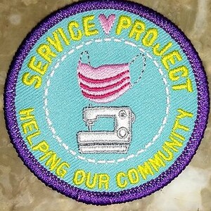 May include: A yellow and purple embroidered patch with the text "Service Project Helping Our Community" and a sewing machine and a pink face mask.