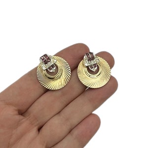 May include: A pair of gold tone earrings with a circular design. The earrings feature a smaller circle with a pink rhinestone band and a larger circle with a textured surface.