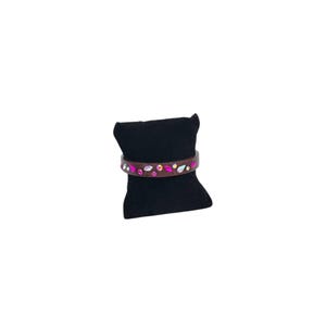 May include: A brown leather bracelet with pink and iridescent gemstones. The gemstones are arranged in a decorative pattern across the front of the bracelet. The bracelet is displayed on a black velvet pillow.