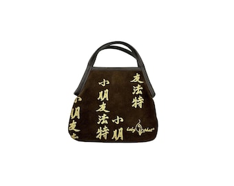 May include: A dark brown suede handbag featuring gold-colored Chinese characters printed across the front surface. The bag has two dark brown handles and includes the text "baby phat" with a cat logo printed in gold on the lower right side.