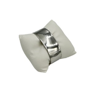 May include: A wide, polished silver cuff bracelet with a curved design. The bracelet is displayed on a white, square pillow. The metal reflects light, creating highlights and shadows.