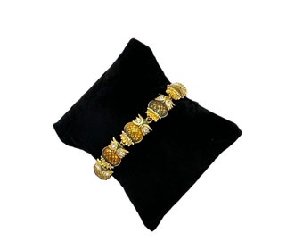 May include: Gold-toned bracelet featuring pineapple-shaped links with green and yellow enamel detailing. The bracelet is adorned with small, clear gemstones and rests on a black velvet surface. A tropical-themed jewelry piece.