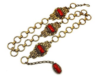 May include: A vintage gold-tone belt with three ornate rectangular coral-colored stone accents. The belt features a chain design with a matching coral-colored stone pendant at the end. The belt is displayed on a white background.