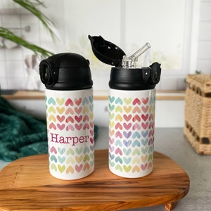 May include: Two white water bottles with black lids and a colorful heart pattern. One bottle has the name "Harper" in pink. The bottles are on a wooden tray. One bottle's lid is open, revealing a straw.
