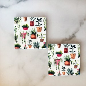 May include: Two square coasters with a white background and a repeating pattern of potted plants in various colors. The plants are in terracotta, pink, and brown pots, with green foliage. The coasters are likely made of ceramic or a similar material.
