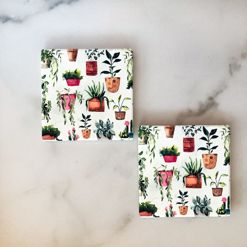 May include: Two square coasters with a white background and a repeating pattern of potted plants in various colors. The plants are in terracotta, pink, and brown pots, with green foliage. The coasters are likely made of ceramic or a similar material.