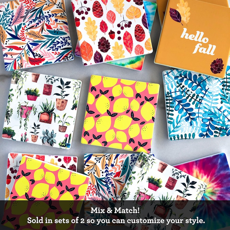 May include: A collection of square ceramic coasters with various designs, including botanical, lemon, and fall-themed patterns. One coaster features the text "hello fall." The coasters are designed to be mixed and matched.