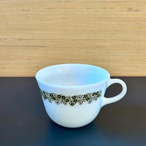 May include: White ceramic teacup with a small handle and a decorative band of green floral pattern around the rim. The cup is empty and sits on a dark surface, with a light-colored wooden background.