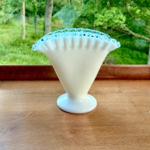 May include: A fan-shaped milk glass vase with a smooth, opaque white body and a flared, ruffled rim. The top edge of the vase features a distinct, vibrant blue glass trim that accents the crimped design of the opening, set on a circular base.