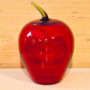 May include: A vibrant red glass apple sculpture with a green stem. The decorative piece has a glossy finish and is set against a light wood-grain background. A unique home decor item.