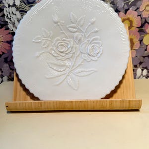 May include: A white, round, vintage cake plate with a scalloped edge. The plate features a raised floral design of roses and leaves. The plate is displayed on a wooden stand against a floral patterned background.