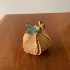 May include: A ceramic pumpkin sculpture with a glossy orange finish and a textured basket-weave pattern. The pumpkin features a curved brown stem and a single light blue leaf attached to the top, showcasing a smooth, reflective glazed surface.