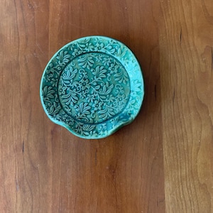 May include: A round, teal-colored ceramic spoon rest featuring a raised, intricate floral and leaf pattern across its entire surface. The dish has a slightly scalloped edge and a glossy finish that highlights the detailed botanical relief design.