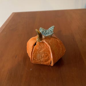 May include: A small, orange ceramic pumpkin figurine with a glossy finish and a textured, embossed surface pattern. It features a dark brown stem and a light blue, butterfly-shaped accent perched on top, adding a distinct decorative element to the piece.