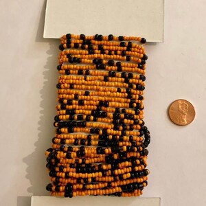 May include: A spool of orange and black seed beads, wrapped in white paper with a small price tag. The beads are small and uniform in size. A UK penny is included for scale.