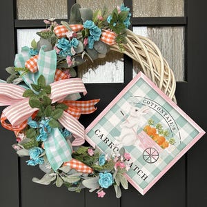 Puede incluir: Una corona decorativa de Pascua con una base tejida, adornada con flores azules y rosas, zanahorias a cuadros naranjas y blancas y un gran lazo a rayas rosas y naranjas. Un letrero dice "Cottontail's Carrot Patch".