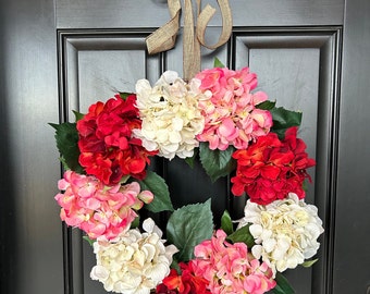 May include: A pink, red, and white flower wreath with green leaves. The wreath is hanging on a dark brown door.
