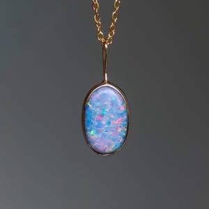 May include: A gold-colored necklace with an oval pendant. The pendant features a blue opal with flashes of pink, green, and yellow. The chain is delicate and hangs from the top of the pendant.
