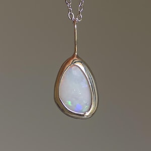 May include: A teardrop-shaped pendant necklace featuring a white opal gemstone with iridescent flashes of color. The opal is set in a polished silver-tone metal frame and hangs from a delicate silver chain. The pendant is approximately 1.5 inches long.