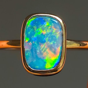 May include: A gold ring featuring a rectangular opal gemstone. The opal displays a vibrant play of color, with hues of blue, green, and orange. The ring's band is a warm gold tone, complementing the gemstone's colors.