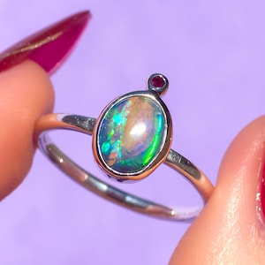 May include: A silver ring featuring an oval opal gemstone, showcasing iridescent green, blue, and yellow hues. A small, round, red gemstone is set above the opal. The ring is set against a purple background.