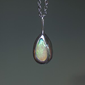 May include: A teardrop-shaped pendant necklace featuring a polished silver-toned metal setting that encases a smooth, iridescent opal gemstone. The opal displays shifting flashes of green and orange light. The pendant hangs from a silver-toned chain.