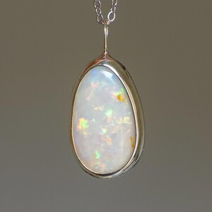 May include: A teardrop-shaped pendant with a large, iridescent opal stone set in a silver bezel. The opal displays flashes of green, orange, and yellow. The pendant hangs from a delicate silver chain.