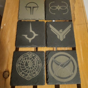 Puede incluir: Seis posavasos cuadrados de pizarra con diseños grabados. Los posavasos presentan varios patrones geométricos y abstractos en tonos grises y beige. Están dispuestos sobre un palé de madera.